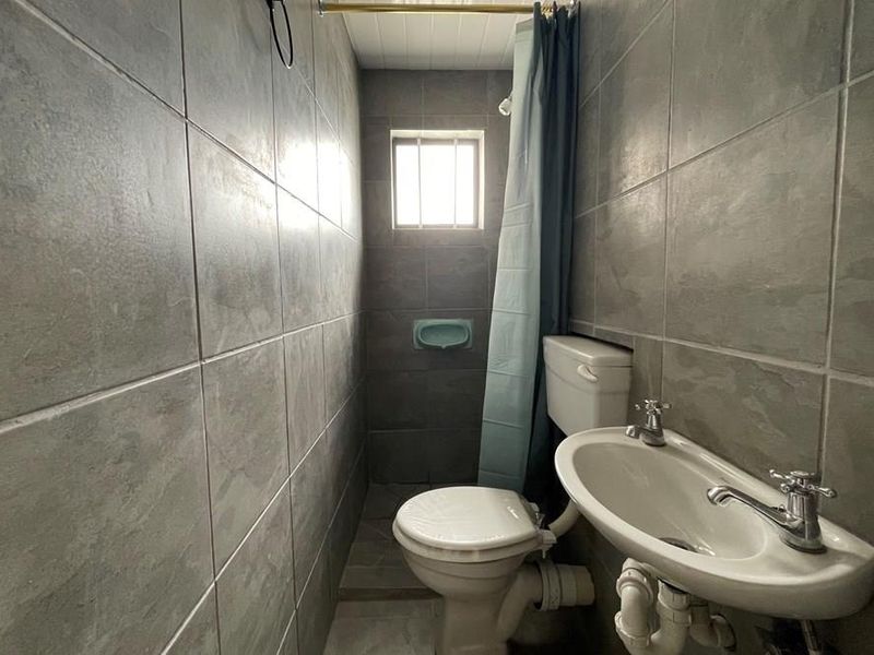 Compact bathroom featuring full-height grey ceramic wall tiling, a pedestal basin, and a toilet, with a small window providing natural light.