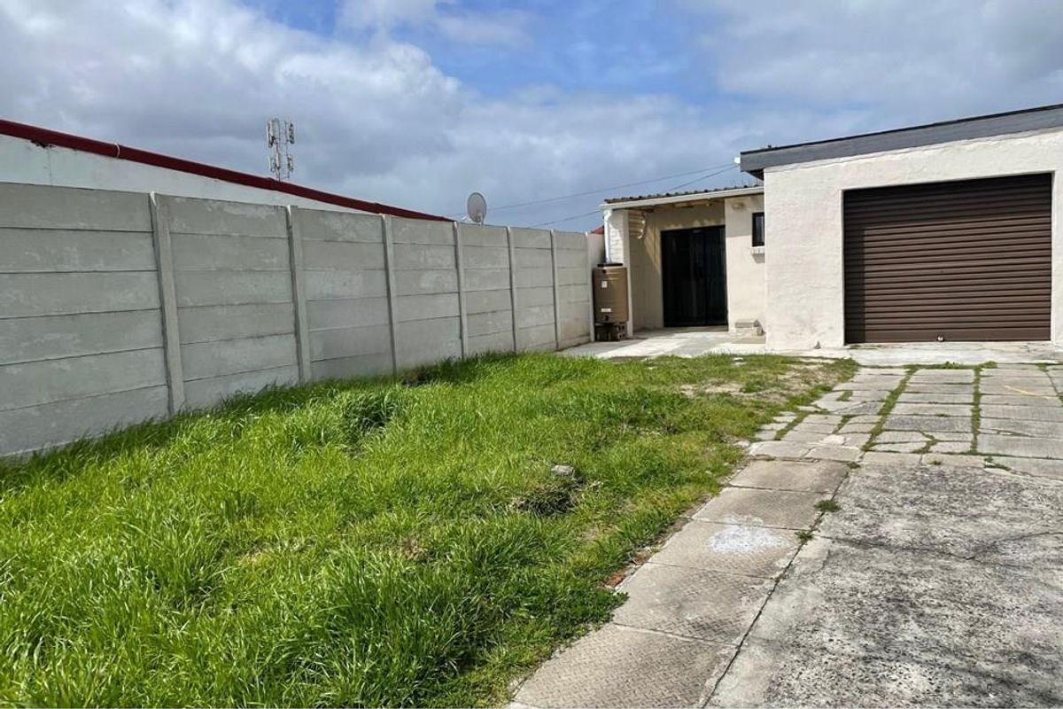 Rear exterior view featuring a paved driveway, roller shutter garage door, and a precast concrete boundary wall with an adjacent lawn area.