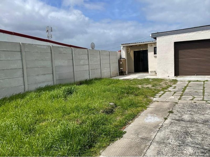 Rear exterior view featuring a paved driveway, roller shutter garage door, and a precast concrete boundary wall with an adjacent lawn area.