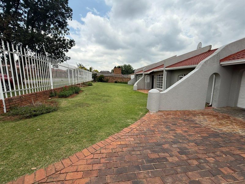 A well-maintained front exterior view featuring a brick driveway, manicured lawn, and secure perimeter fencing with a tiled roof facade.