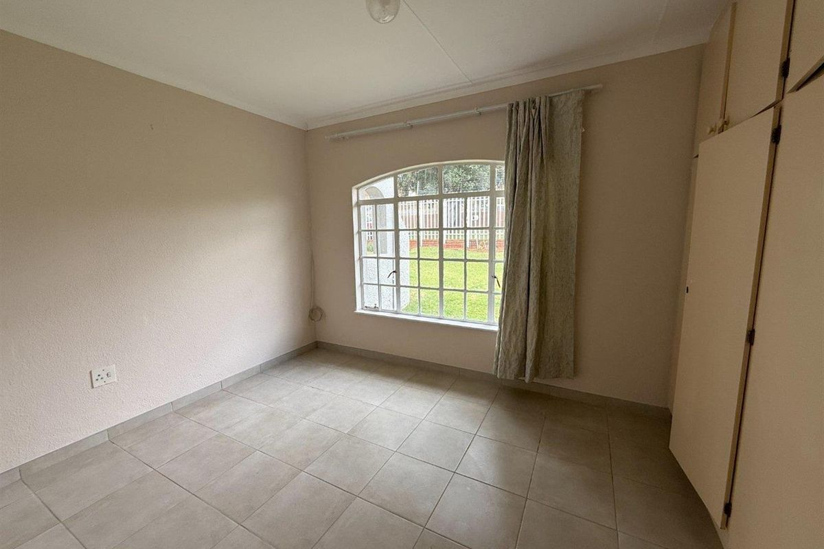 A bright, empty bedroom featuring neutral tiled flooring, built-in cupboards, and an arched window overlooking the garden.