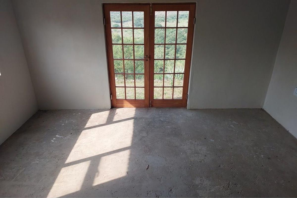 An empty, unfinished room featuring raw concrete flooring and wooden French doors that open to a scenic landscape view.
