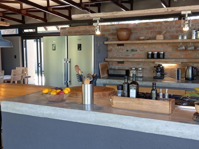 Spacious open-plan kitchen featuring a large concrete island, exposed brick wall, timber shelving, and integrated oven, seamlessly flowing into the dining area.