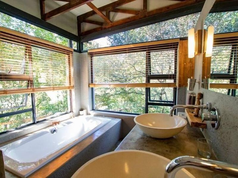 A modern bathroom featuring dual vessel sinks, a built-in bathtub, exposed timber roof trusses, and large windows offering scenic views.