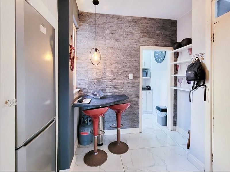 A compact kitchen area featuring a stone-clad feature wall, built-in breakfast bar, modern pendant lighting, and polished tiled flooring, leading into a secondary kitchen space.