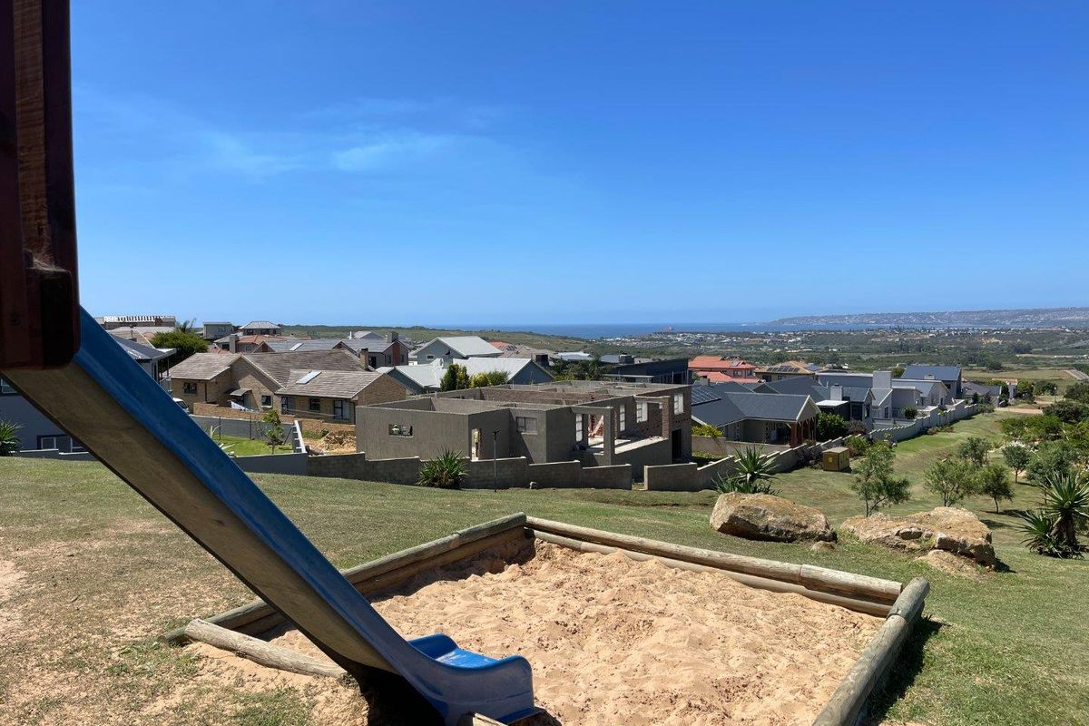 Elevated view overlooking a residential neighbourhood and distant coastline, featuring a landscaped garden area with a sandpit and slide.