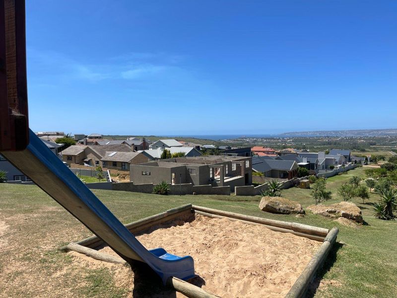 Elevated view overlooking a residential neighbourhood and distant coastline, featuring a landscaped garden area with a sandpit and slide.