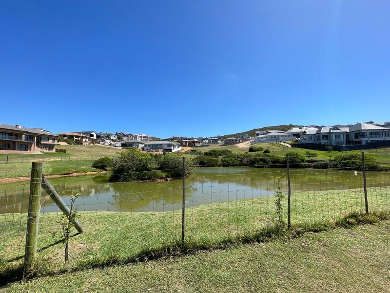 A scenic view of a residential estate featuring a central pond, landscaped green areas, and surrounding modern homes under a clear blue sky.