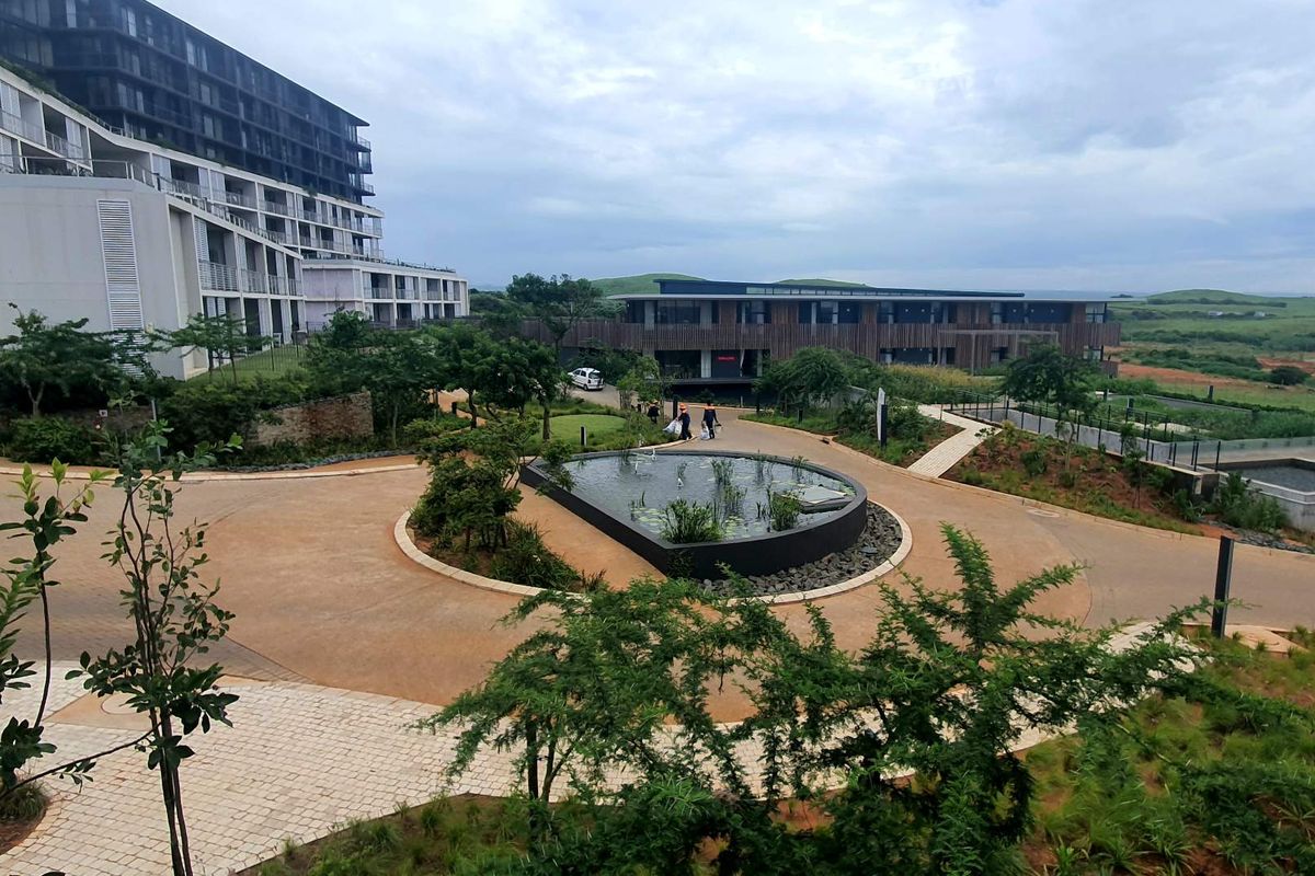 An elevated view of the apartment complex grounds, featuring a central circular water feature, landscaped gardens, paved pathways, and modern multi-story residential buildings.