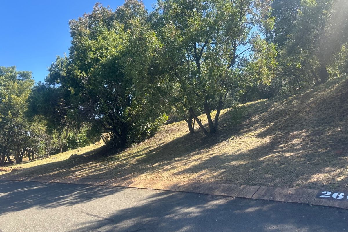 A street-level view of a sloped residential vacant land plot featuring mature trees and established road infrastructure.