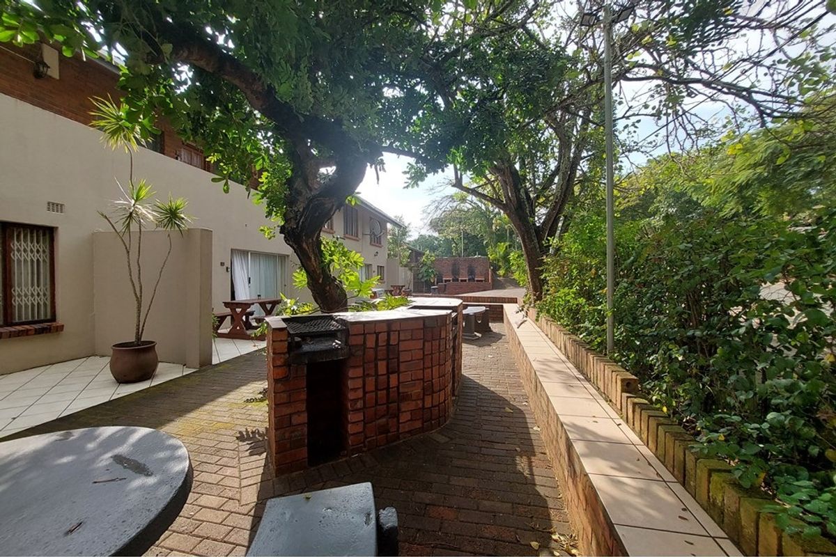 Outdoor communal entertainment area featuring a built-in brick braai, paved patio space, and established trees within a residential apartment complex setting.