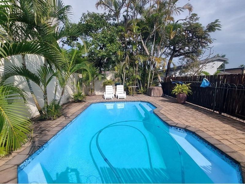 A well-maintained outdoor swimming pool area featuring a paved deck, perimeter fencing, and established tropical landscaping.
