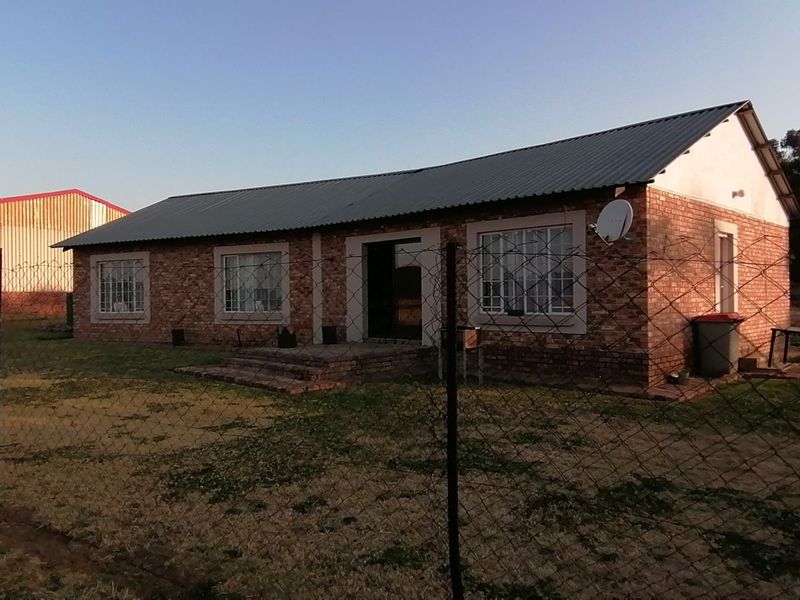 A single-story brick farmhouse featuring a metal roof, security fencing, and a large adjacent agricultural shed on a rural property.