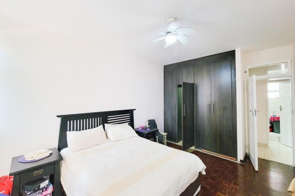 A well-maintained bedroom featuring dark wood built-in cupboards, a ceiling fan, and classic parquet flooring, with an adjoining doorway leading to another room.