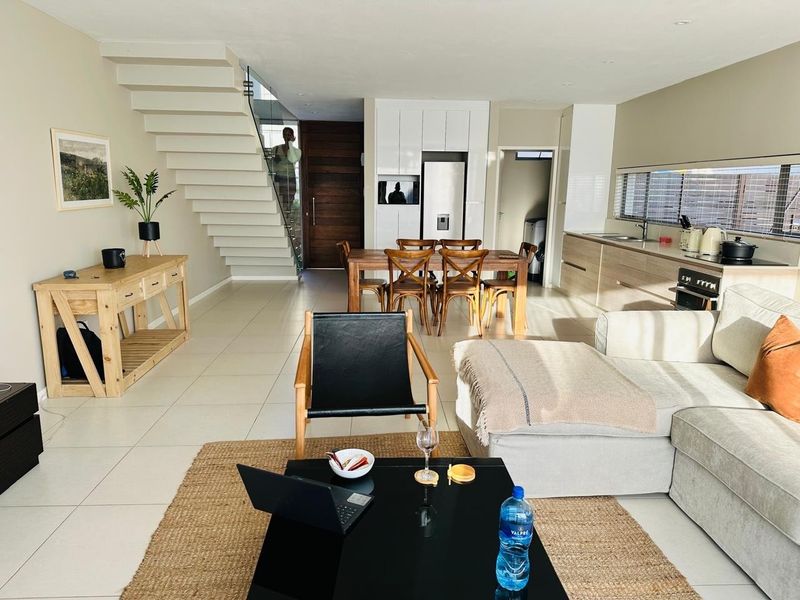 Spacious open-plan living, dining, and kitchen area featuring modern cabinetry, a sleek staircase with glass balustrade, and neutral tiled flooring throughout.