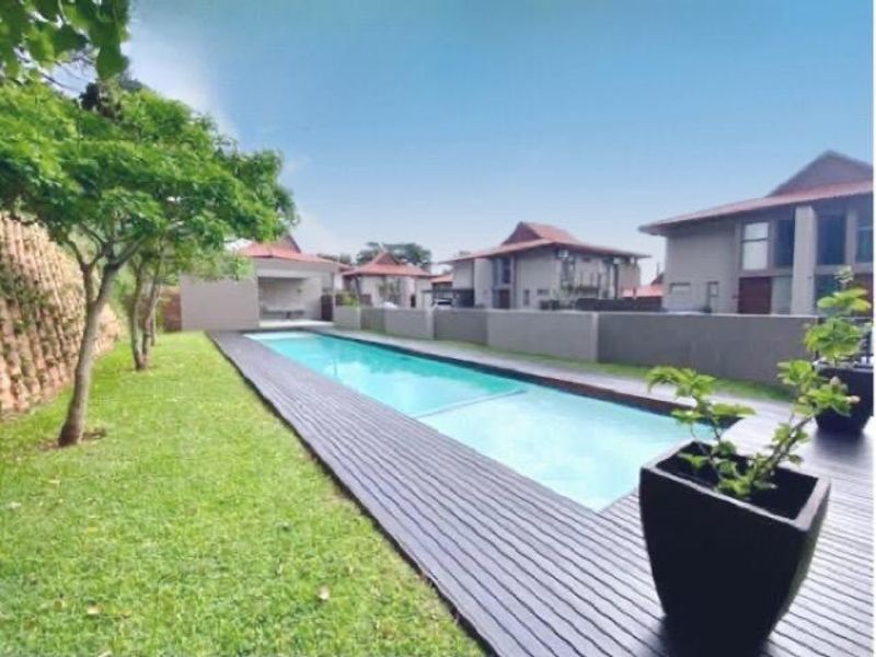 A well-maintained outdoor pool area featuring a long swimming pool, timber decking, and a landscaped garden with a retaining wall.