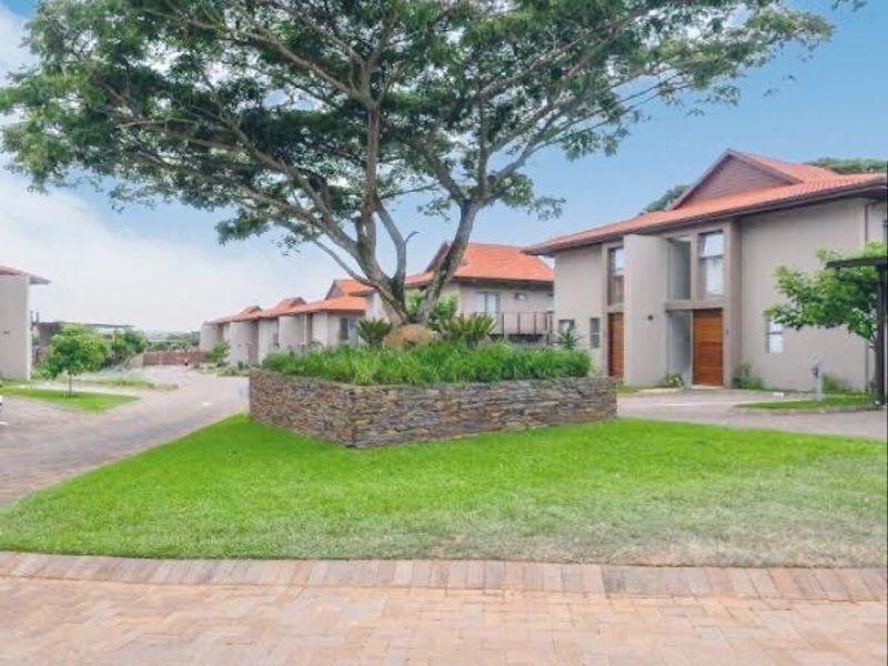 A view of the townhouse complex entrance featuring a landscaped stone retaining wall, paved driveway, and modern residential units with tiled roofs.