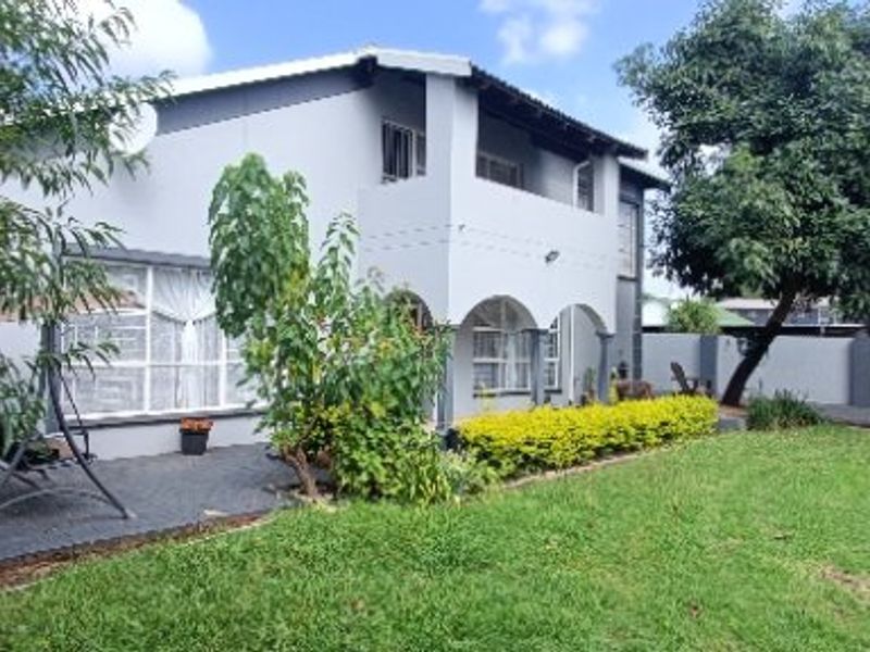 Rear view of a two-story house featuring a paved patio, arched veranda, upper-level balcony, and a well-maintained garden with perimeter walling.