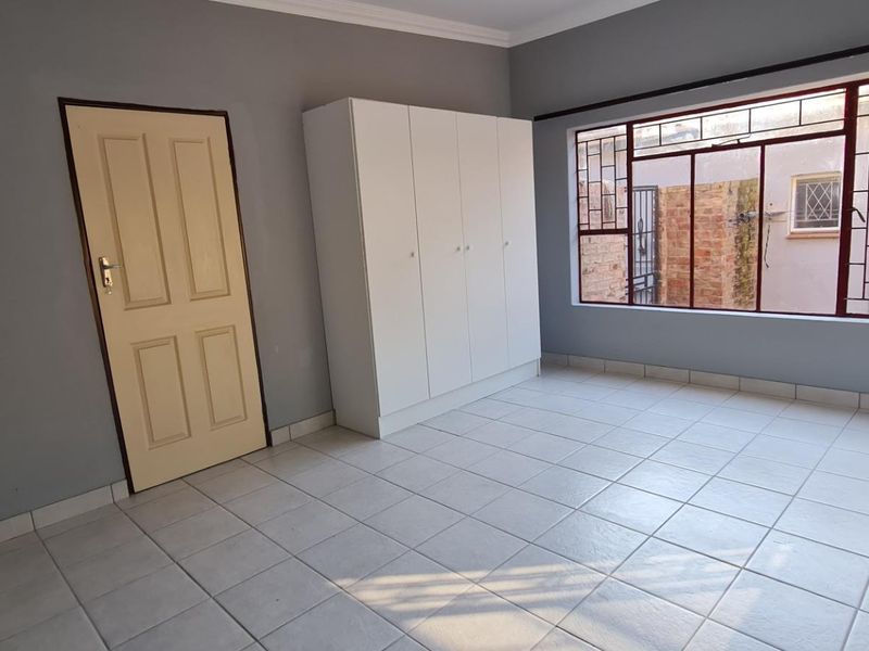 A bright, empty bedroom featuring neutral grey walls, tiled flooring, and a built-in white wardrobe with security bars on the window.