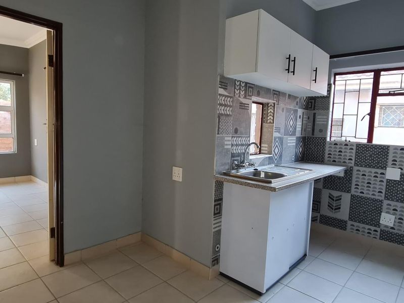A compact kitchen featuring patterned tiled splashbacks, stainless steel sink, and built-in cabinetry, leading into an adjacent room with tiled flooring.
