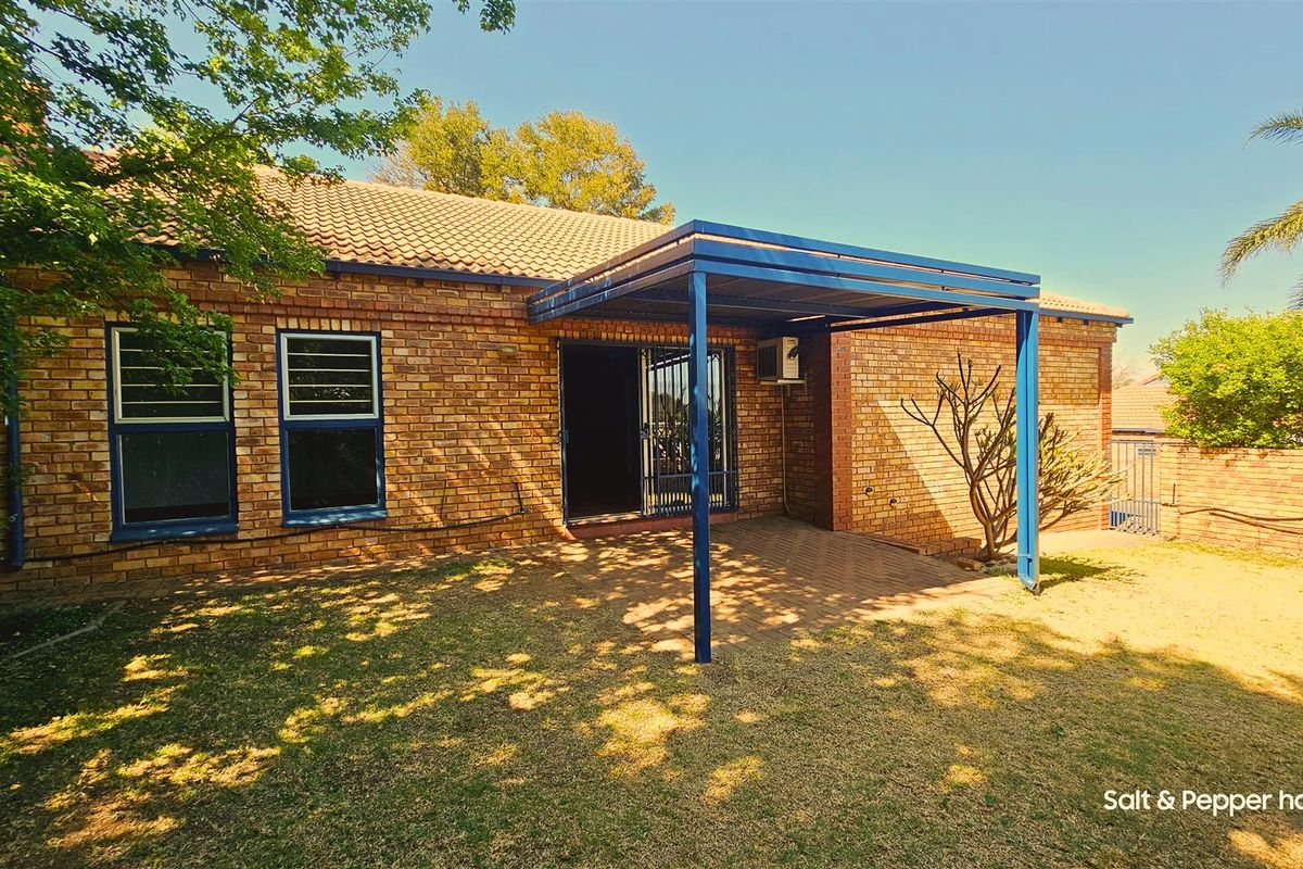 Rear view of the townhouse featuring a brick facade, a paved patio area covered by a blue metal pergola, and a small garden.