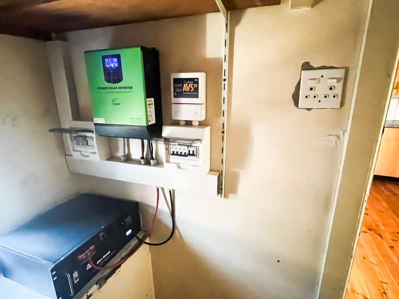 Utility area featuring a wall-mounted hybrid solar inverter, voltage protection device, and electrical distribution board with visible wall surface damage.