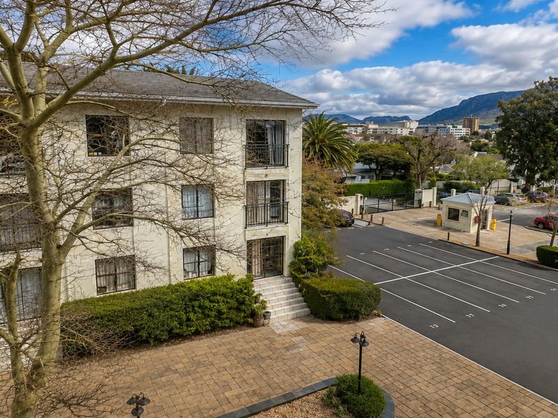 Elevated view of the apartment complex exterior, featuring a paved courtyard, designated parking bays, and a security gatehouse with mountain views in the background.