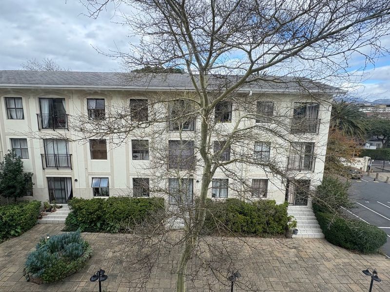 A view of the apartment building's rear facade, featuring a paved courtyard, landscaped garden beds, and balconies with wrought iron railings.