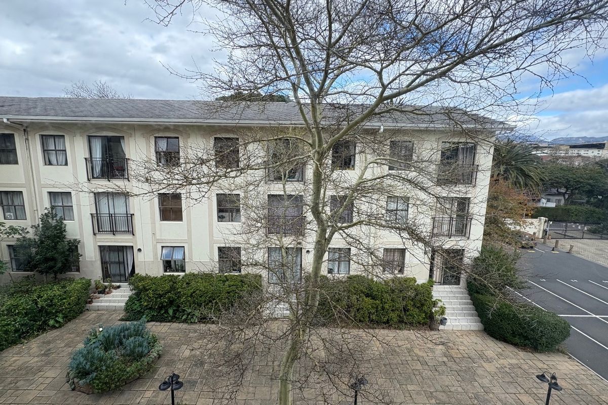 A view of the apartment building's rear facade, featuring a paved courtyard, landscaped garden beds, and balconies with wrought iron railings.