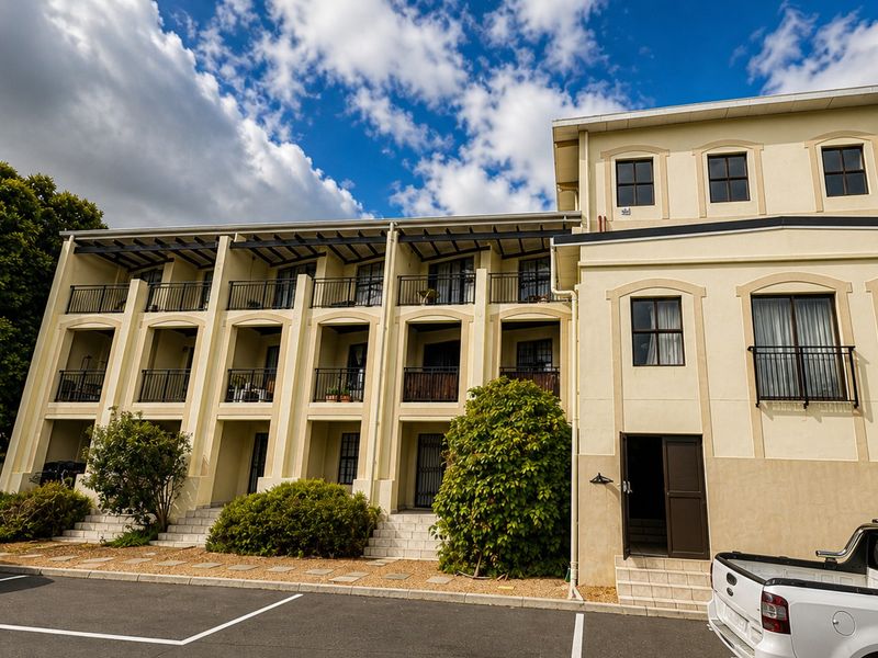 Front elevation of a multi-story apartment building featuring private balconies, metal railings, and ground-level access points with paved parking.