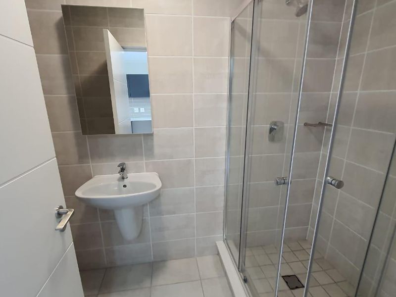A modern, clean bathroom featuring a pedestal washbasin, wall-mounted mirror, and a glass-enclosed shower with neutral ceramic tiling throughout.