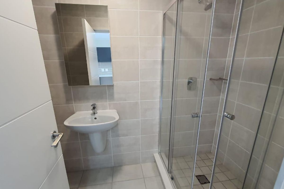 A modern, clean bathroom featuring a pedestal washbasin, wall-mounted mirror, and a glass-enclosed shower with neutral ceramic tiling throughout.