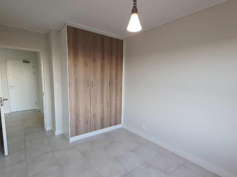 A clean, modern bedroom featuring neutral tiled flooring, built-in wood-finish cupboards, and a simple pendant light fixture.