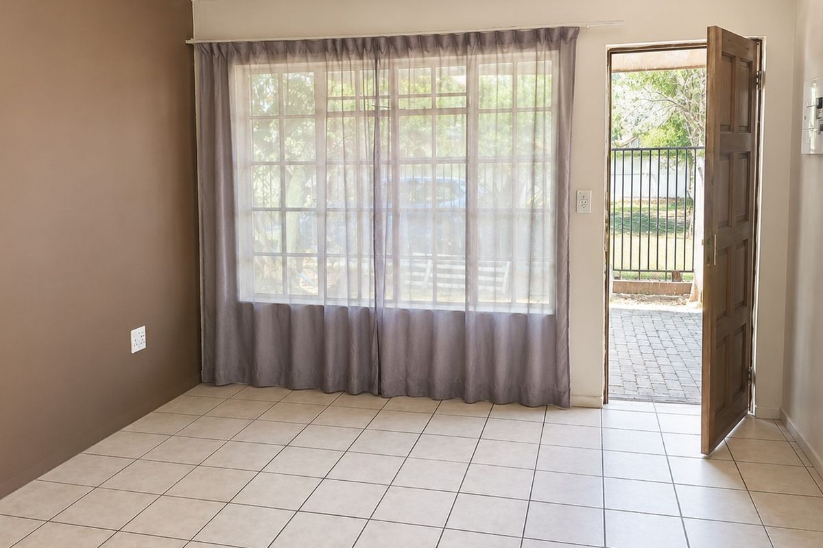 A bright, empty living area featuring tiled flooring, a large grid-pattern window, and direct access to an outdoor patio space.