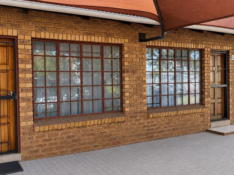 Front exterior view of a face brick flat featuring secure timber doors, grid windows, and a paved entrance area under a shade sail.