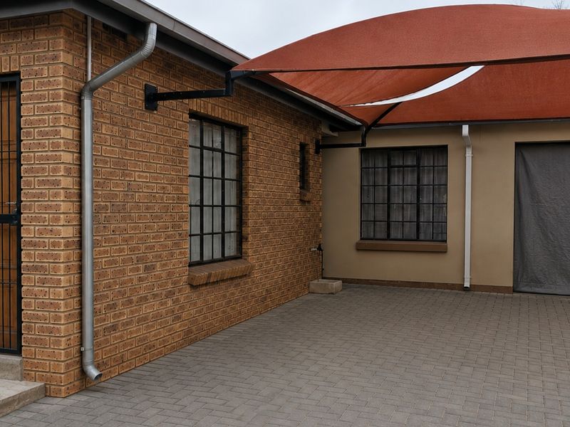 A paved rear courtyard area featuring brick exterior walls, steel-framed windows, a security gate, and a large overhead shade sail.