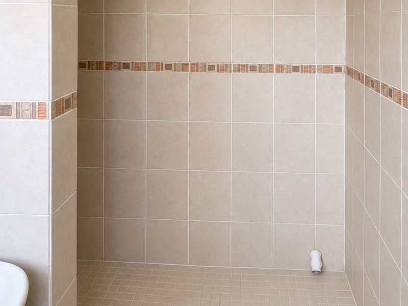 A clean, tiled shower enclosure featuring neutral ceramic wall tiles with a decorative mosaic border and a dedicated plumbing outlet.
