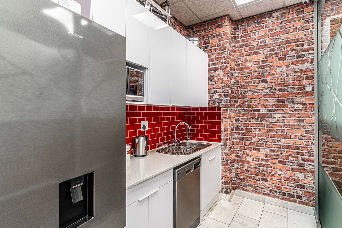 A modern office kitchenette featuring white cabinetry, a striking red subway tile backsplash, exposed brick wall, and integrated appliances.