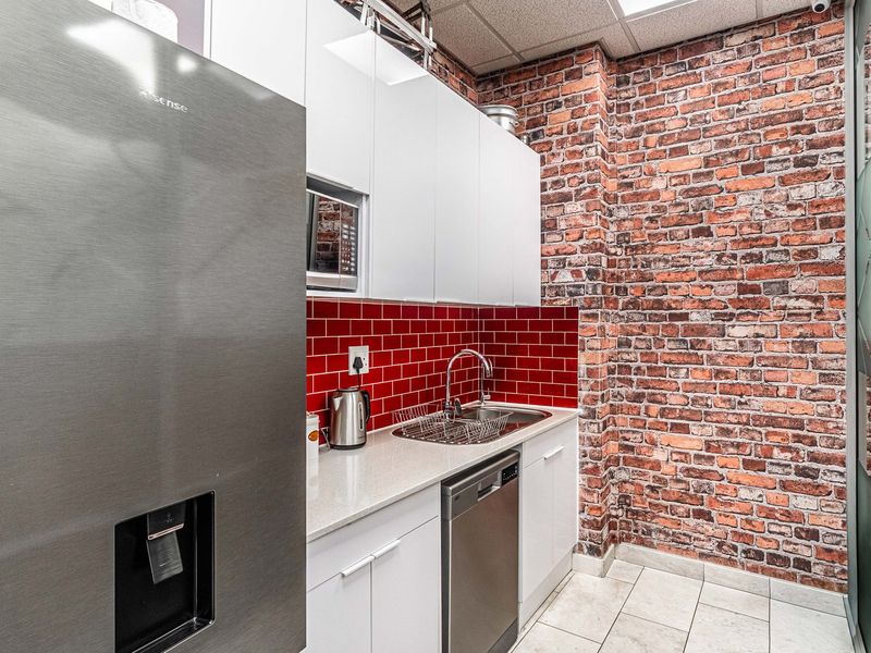 A modern office kitchenette featuring white cabinetry, a striking red subway tile backsplash, exposed brick wall, and integrated appliances.