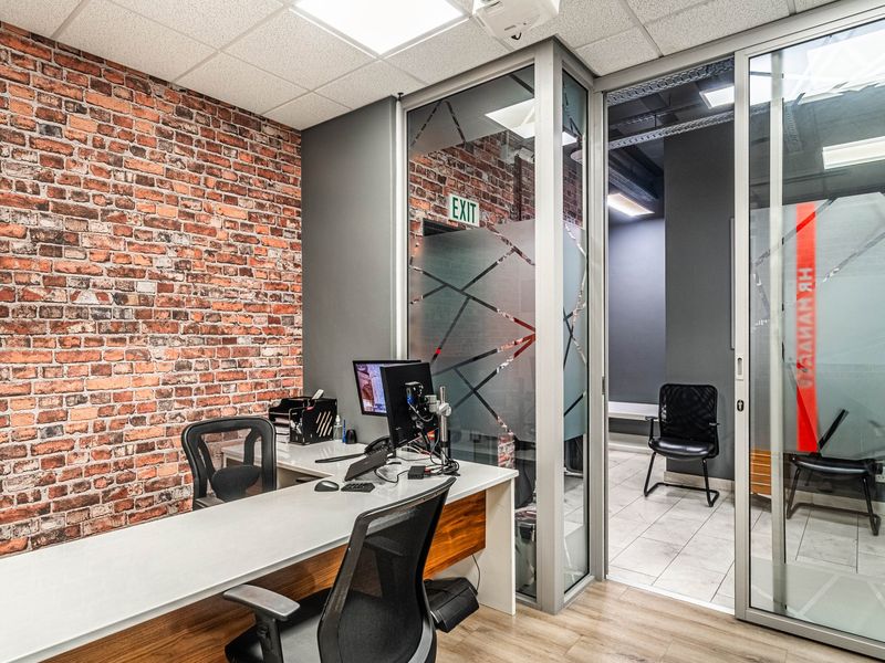 Modern office space featuring an exposed brick wall, glass partition walls with privacy film, and contemporary laminate flooring.