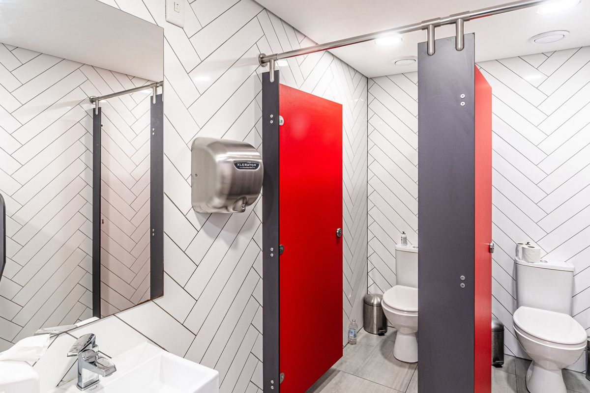 Modern office bathroom featuring herringbone wall tiling, partitioned toilet stalls, wall-mounted hand dryer, and clean, contemporary fixtures.