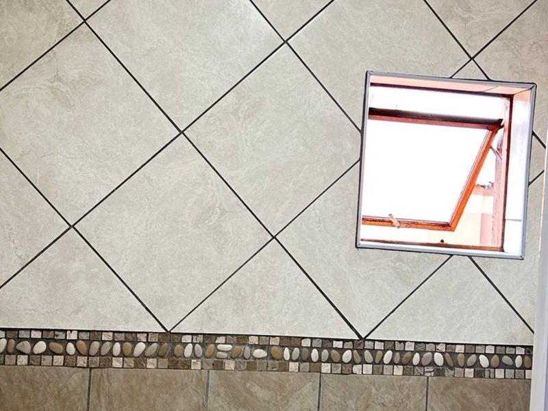 A bathroom wall featuring neutral diamond-patterned tiles with a decorative mosaic border and a functional top-hung window.
