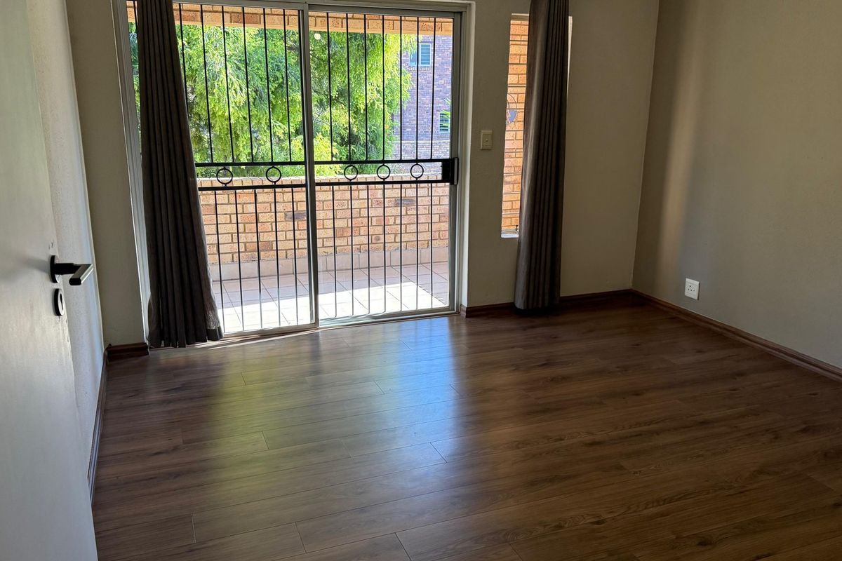A bright, empty bedroom featuring modern laminate flooring and a sliding glass door with an integrated security gate leading to an outdoor balcony.