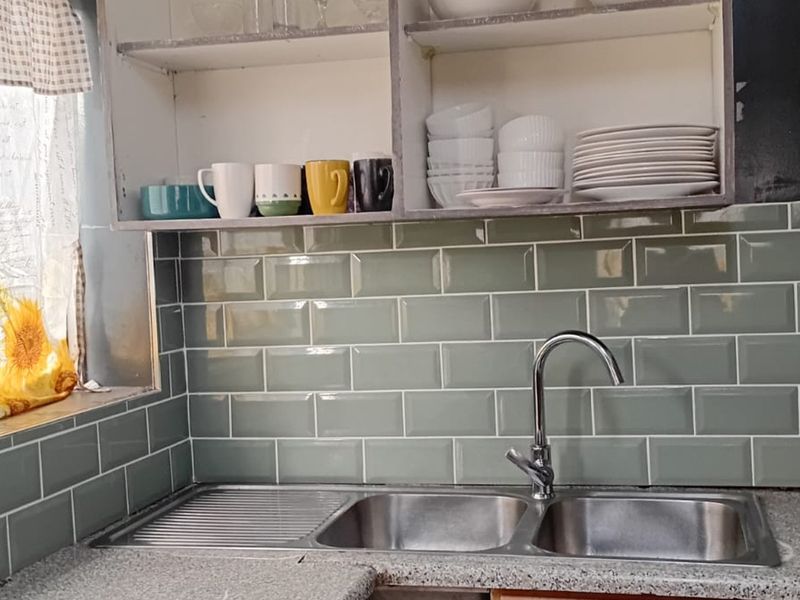 A functional kitchen area featuring a modern sage green subway tile backsplash, a double bowl stainless steel sink, and open shelving storage.