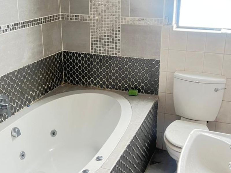 A functional bathroom featuring a built-in jetted bathtub, pedestal basin, and toilet, finished with decorative grey and black mosaic wall tiling.
