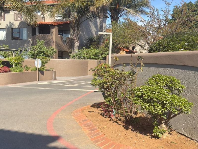 View of the townhouse complex entrance featuring an asphalt driveway, perimeter boundary wall, and landscaped garden beds.