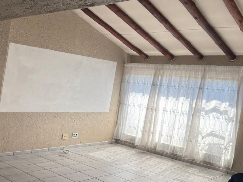 An empty living room featuring exposed timber ceiling beams, tiled flooring, and textured walls with a recent patch repair.