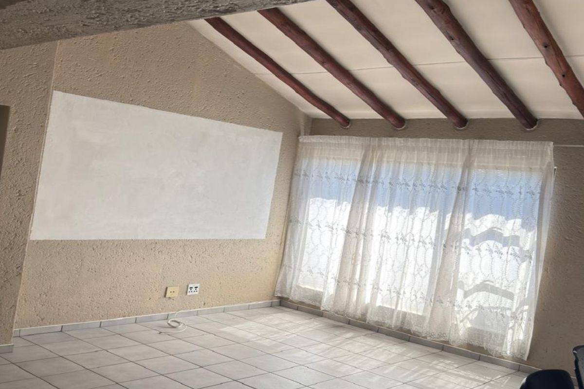 An empty living room featuring exposed timber ceiling beams, tiled flooring, and textured walls with a recent patch repair.