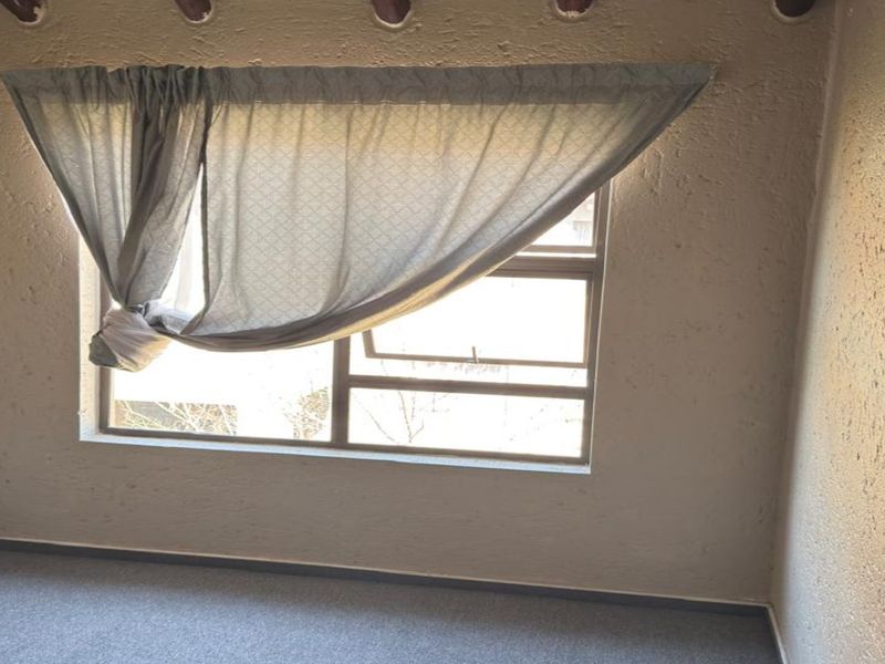 An empty bedroom featuring textured plaster walls, exposed timber ceiling beams, and carpeted flooring with a large window providing natural light.