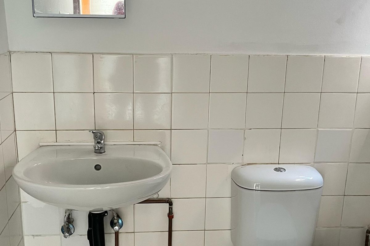 A functional bathroom featuring a pedestal washbasin, toilet, and white ceramic wall tiles with exposed plumbing.